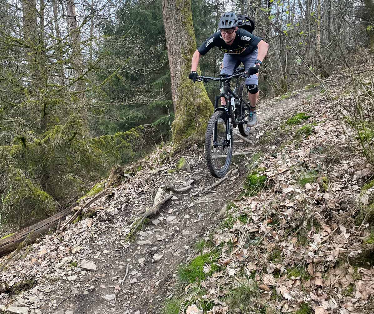 Mountain biker rides a narrow rocky trail in the forest with roots, stones and elevation changes.